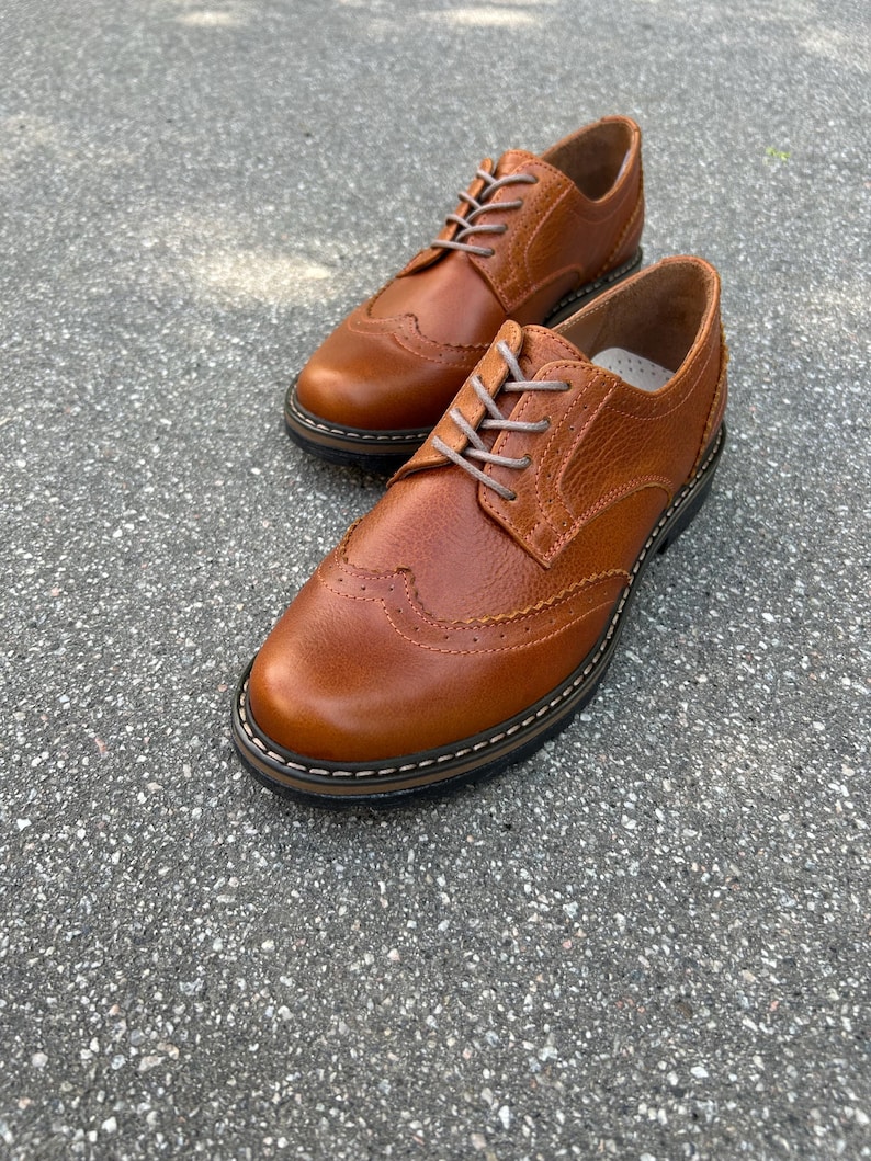 Pu&ograve; includere: Un paio di scarpe Oxford in pelle marrone con perforazioni decorative e lacci colorati chiari. Le scarpe hanno un classico design a punta alata e una suola robusta.