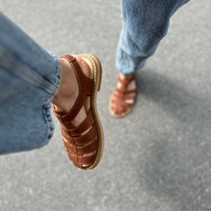 K&ouml;nnte beinhalten: Ein Paar braune Ledersandalen mit geflochtenem Design. Die Sandalen haben eine dicke Sohle und einen Riemen, der um den Kn&ouml;chel geht.