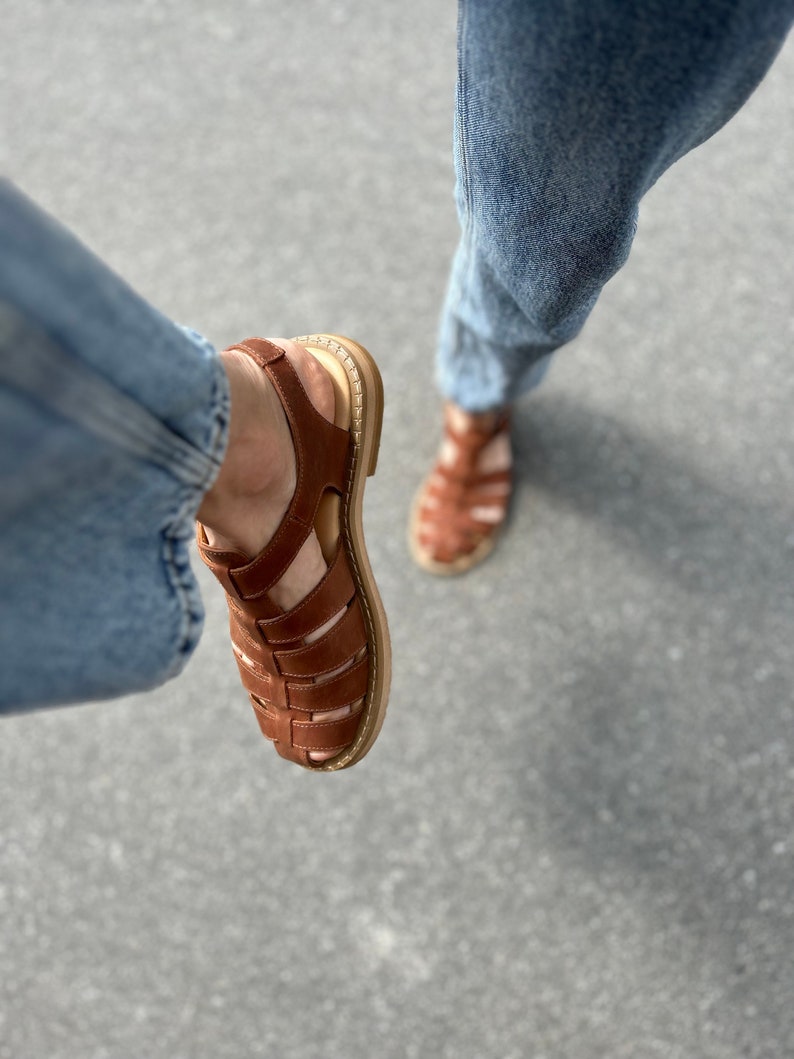 K&ouml;nnte beinhalten: Ein Paar braune Ledersandalen mit geflochtenem Design. Die Sandalen haben eine dicke Sohle und einen Riemen, der um den Kn&ouml;chel geht.