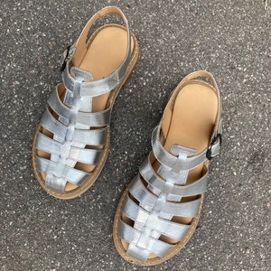 May include: A pair of silver gladiator sandals with a woven design. The sandals have a buckle closure and a flat sole.
