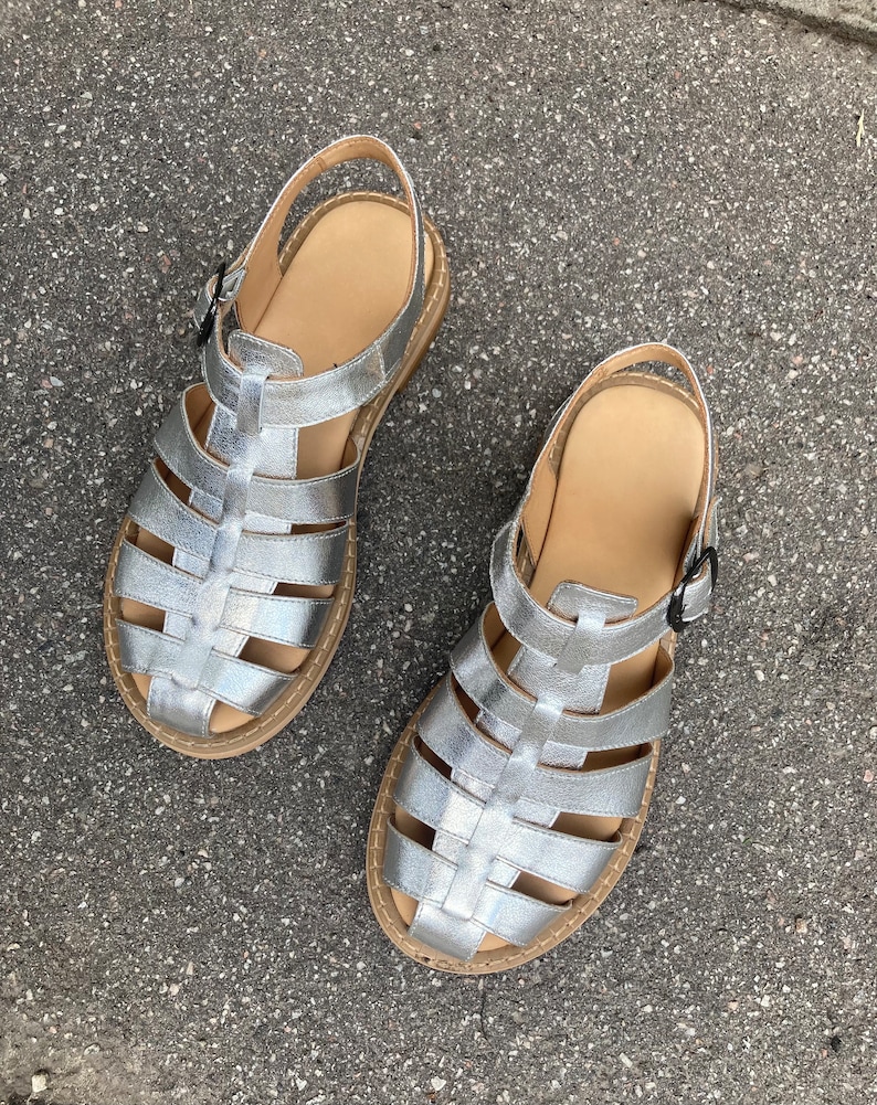 May include: A pair of silver gladiator sandals with a woven design. The sandals have a buckle closure and a flat sole.