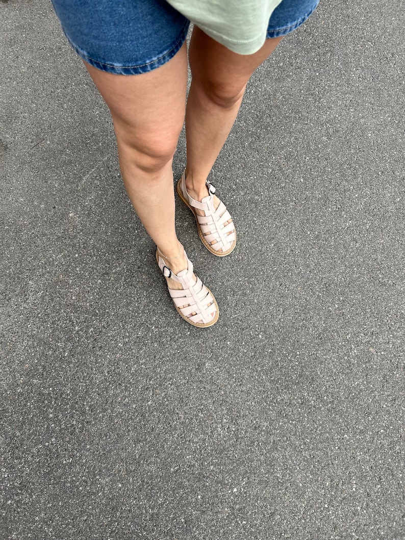 May include: A pair of light pink leather sandals with a caged design and buckle closures. The sandals are worn on a person's feet, standing on a gray asphalt surface.