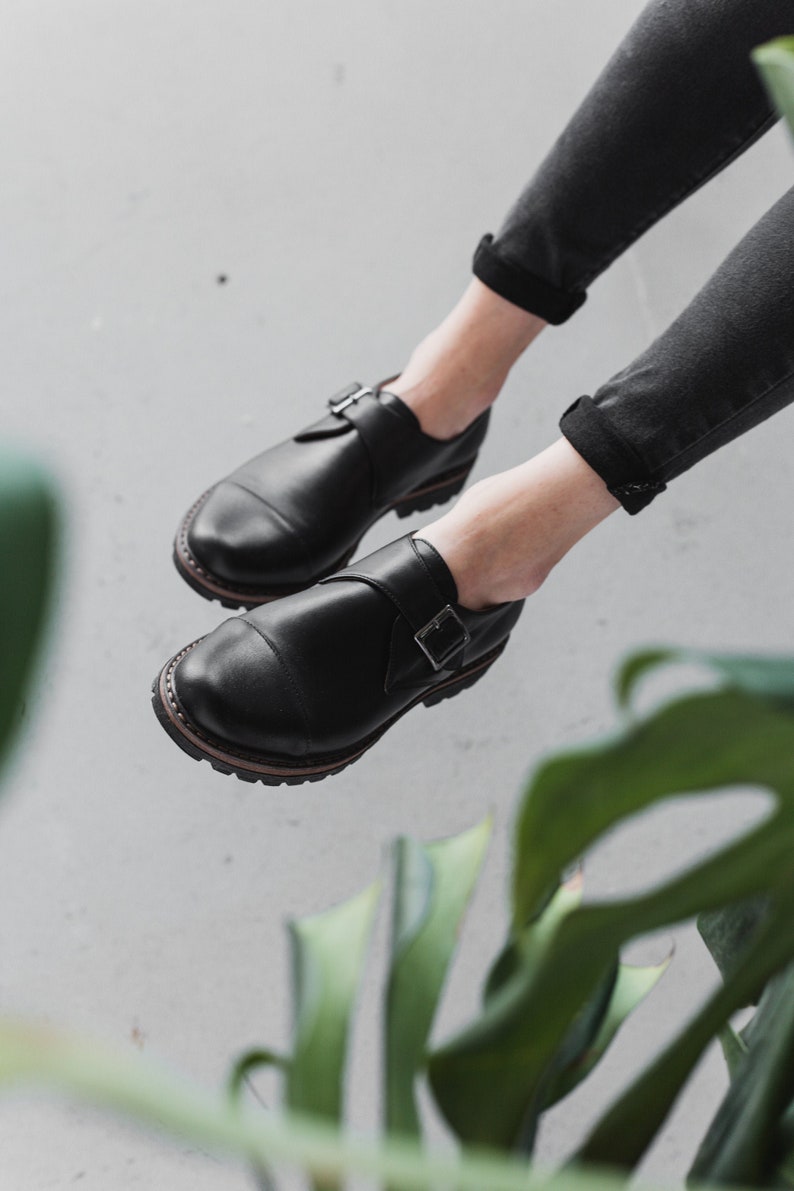 May include: A pair of black leather buckle shoes with a chunky sole. The shoes are worn by a person wearing black jeans.