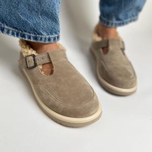 Puede incluir: Un par de zapatos de piel de ante marr&oacute;n claro de deslizamiento con una correa de hebilla y una suela de goma blanca. Los zapatos tienen un forro peludo.