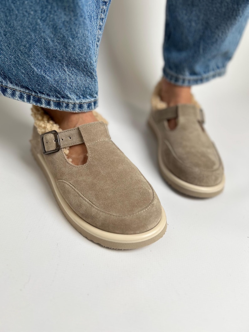 Puede incluir: Un par de zapatos de piel de ante marr&oacute;n claro de deslizamiento con una correa de hebilla y una suela de goma blanca. Los zapatos tienen un forro peludo.