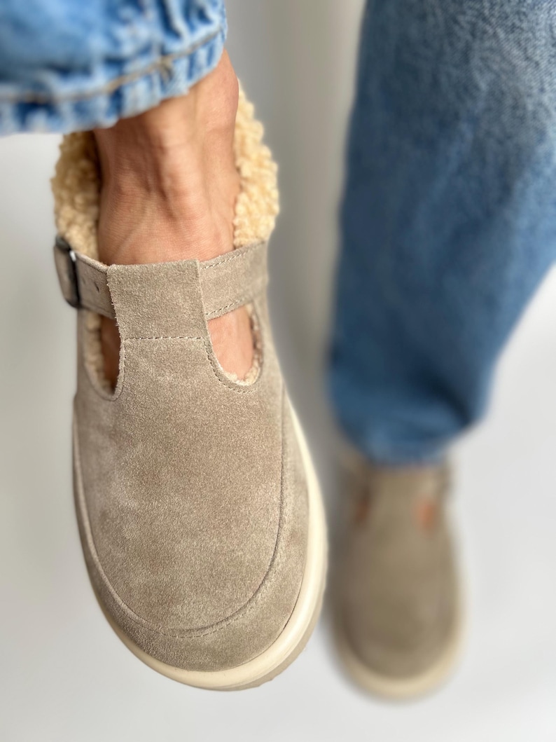 Puede incluir: Un par de zapatos de piel de ante marr&oacute;n claro de deslizamiento con una suela blanca y un forro de piel de oveja. Los zapatos son usados por una persona que lleva pantalones vaqueros azules.