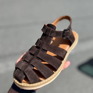 Pu&ograve; includere: Sandali in pelle scamosciata marrone con design intrecciato. I sandali hanno un cinturino con chiusura a fibbia e una suola colorata chiara. Adatti per il clima caldo e l'abbigliamento casual.