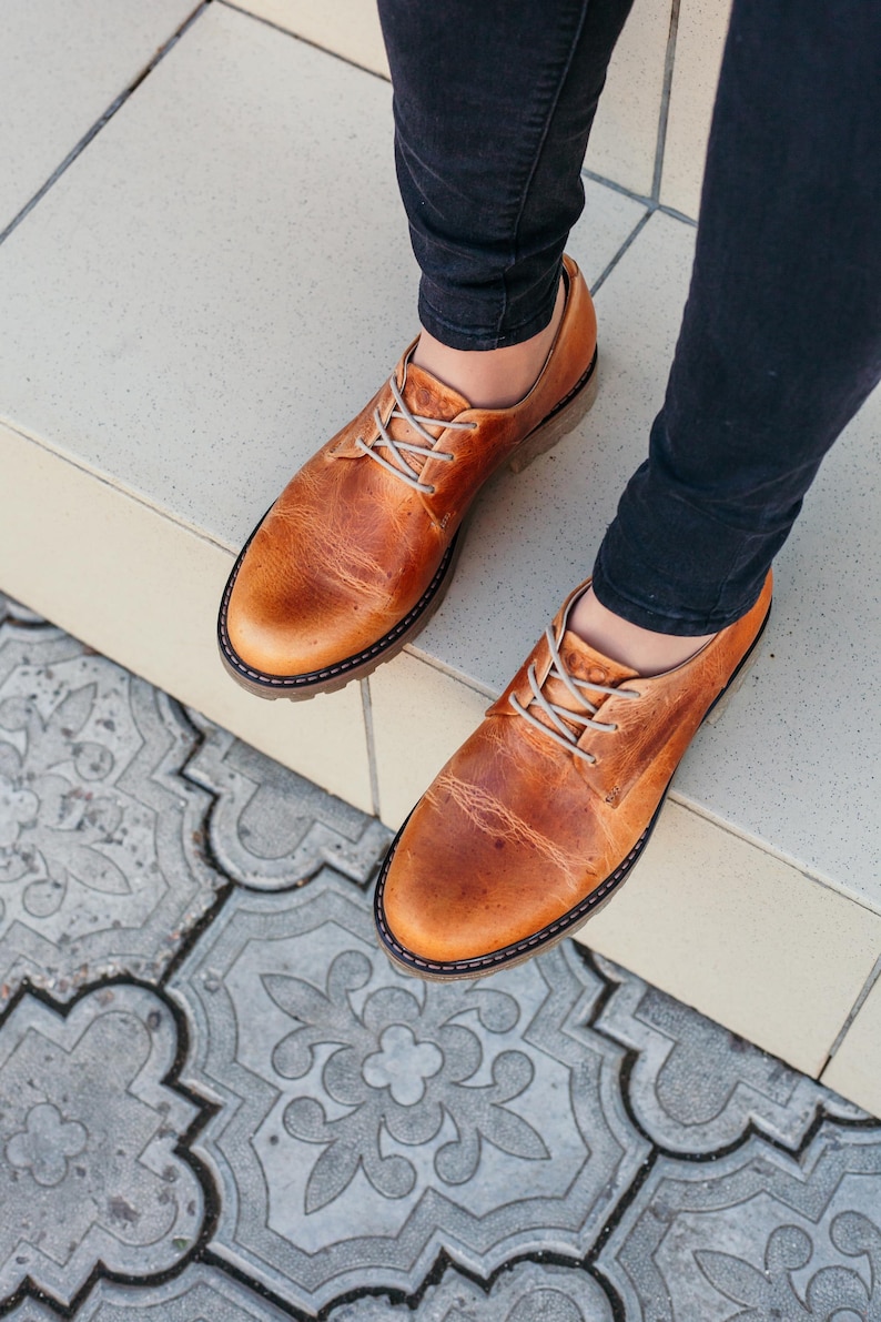 May include: A pair of brown leather lace-up shoes with a slightly distressed look. The shoes are worn by a person wearing dark blue jeans.