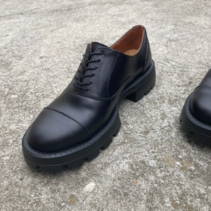May include: Black leather lace-up shoes with a chunky rubber sole. The shoes have a cap toe design and a slightly rounded toe.