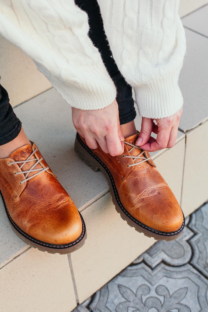 May include: A pair of brown leather lace-up shoes with thick soles. The shoes are being tied by a person wearing a white sweater.