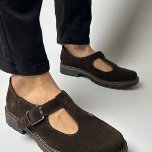 Puede incluir: Zapatos marrones de ante con cierre de hebilla. Los zapatos tienen una puntera redonda y una suela gruesa y texturizada. Se llevan con pantalones negros.