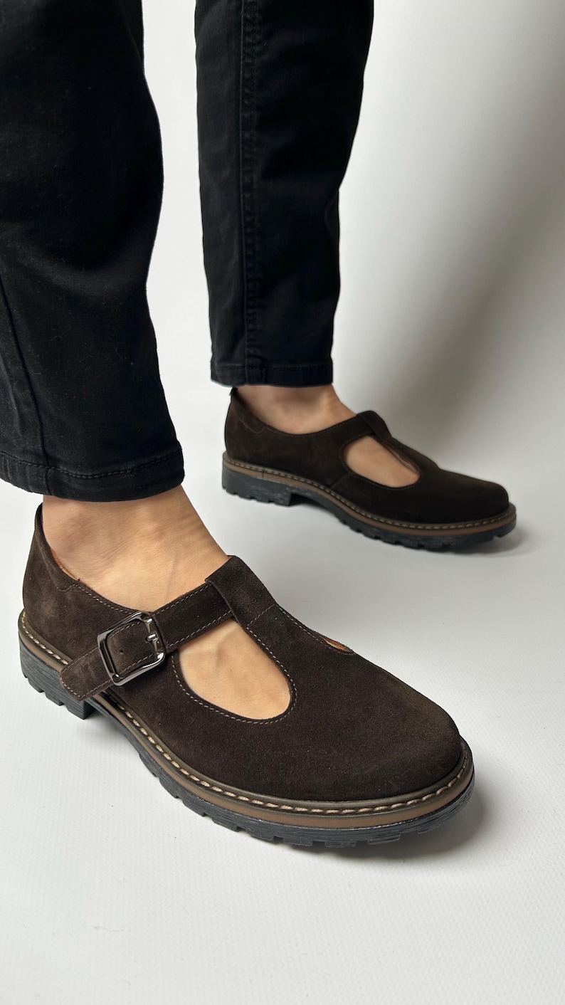 Puede incluir: Zapatos marrones de ante con cierre de hebilla. Los zapatos tienen una puntera redonda y una suela gruesa y texturizada. Se llevan con pantalones negros.