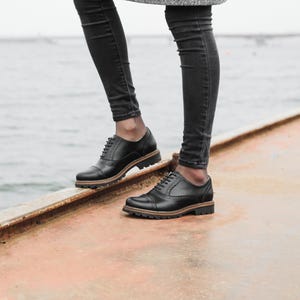 May include: A person wearing black jeans and a gray coat is standing on a metal surface. They are wearing black leather lace-up shoes with a brown sole.