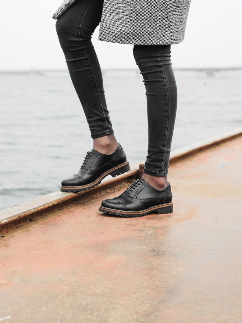 May include: A person wearing black jeans and a gray coat is standing on a metal surface. They are wearing black leather lace-up shoes with a brown sole.
