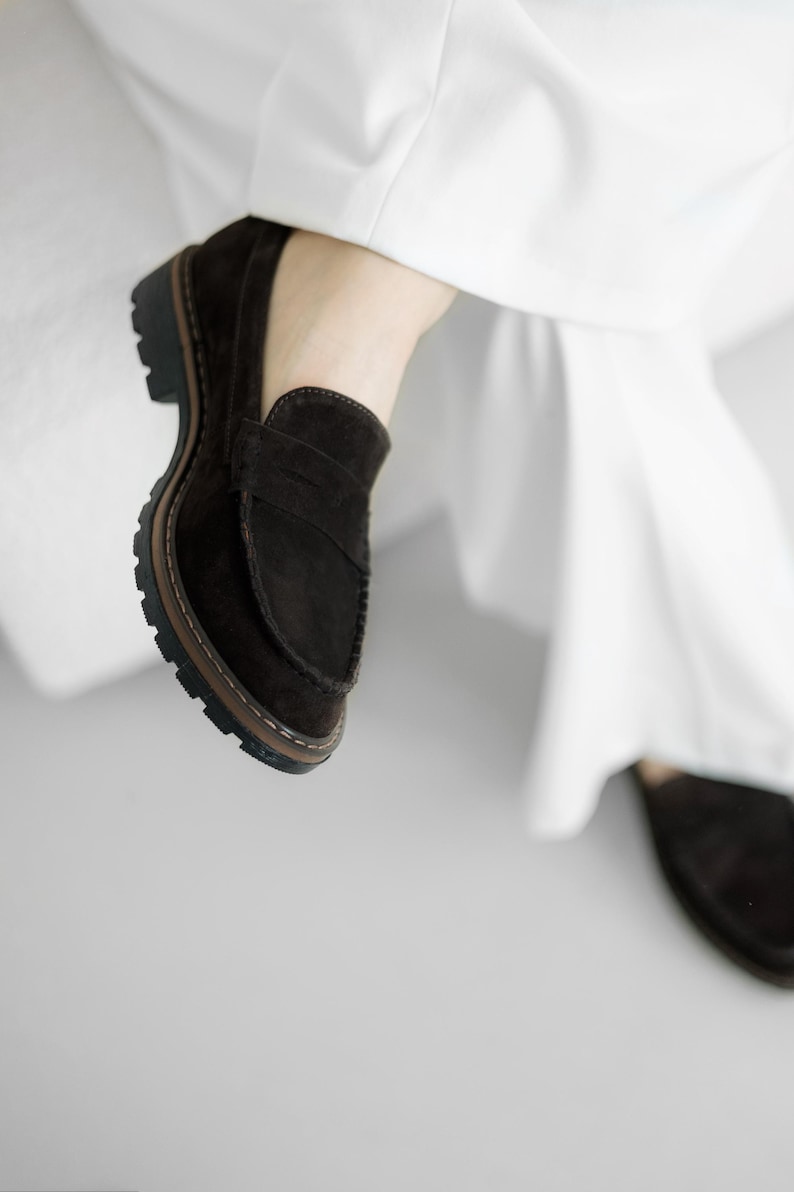 May include: A pair of brown suede loafers with a thick black sole. The loafers have a simple design with a single strap across the top.