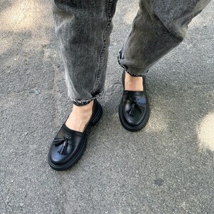 May include: Black leather loafers with a tassel detail. The loafers have a chunky sole and are worn with grey jeans.