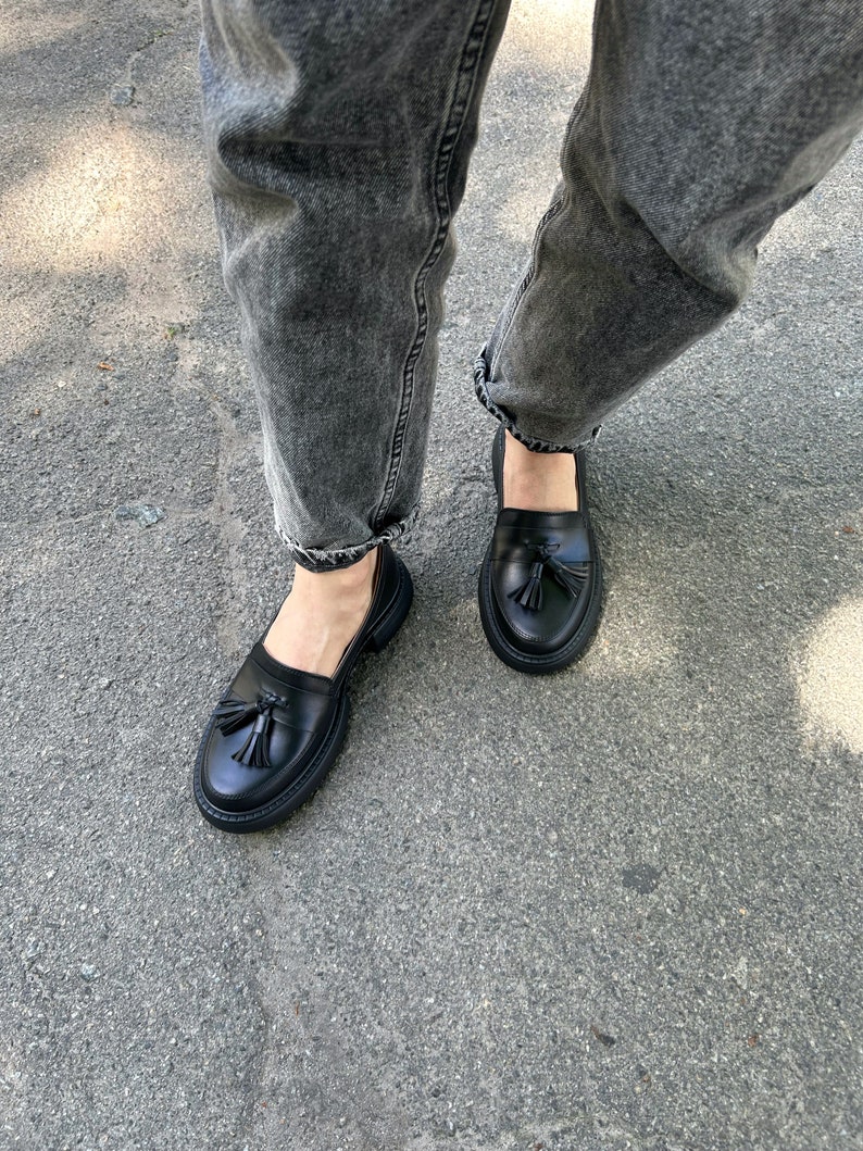 May include: Black leather loafers with a tassel detail. The loafers have a chunky sole and are worn with grey jeans.