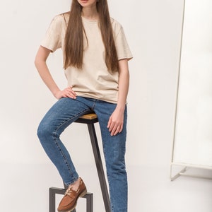 Op de afbeelding: Een vrouw in een beige t-shirt en blauwe jeans zit op een zwarte metalen kruk met een houten zitting. Ze draagt ​​bruine leren bootschoenen.