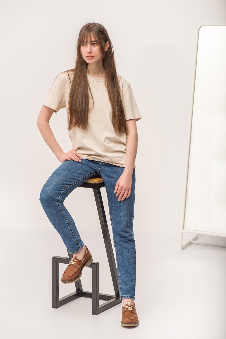 Op de afbeelding: Een vrouw in een beige t-shirt en blauwe jeans zit op een zwarte metalen kruk met een houten zitting. Ze draagt ​​bruine leren bootschoenen.