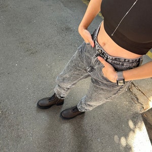 May include: A person wearing a black crop top and light grey acid wash jeans with brown lace-up boots. The person is wearing a silver watch on their left wrist.