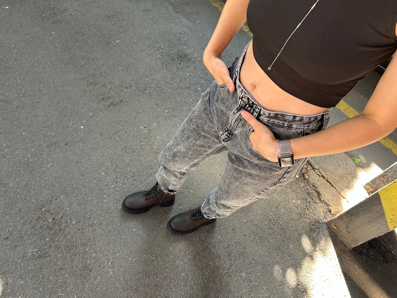 May include: A person wearing a black crop top and light grey acid wash jeans with brown lace-up boots. The person is wearing a silver watch on their left wrist.
