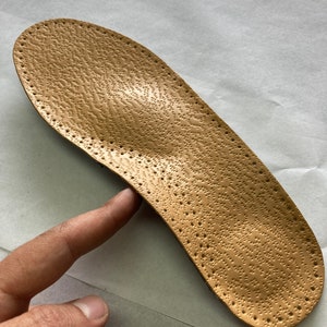 May include: A tan leather insole with a textured surface and small holes throughout. The insole is designed to provide support and comfort for the feet.
