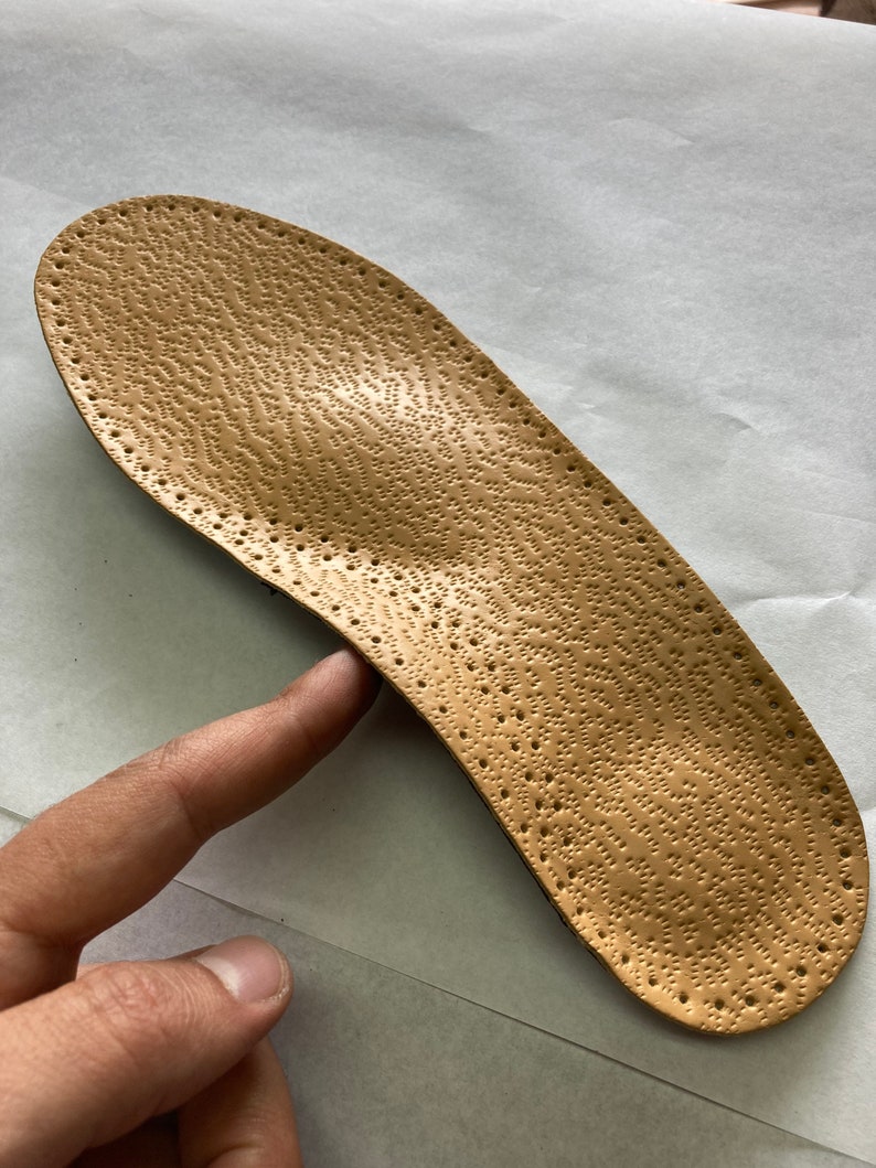 May include: A tan leather insole with a textured surface and small holes throughout. The insole is designed to provide support and comfort for the feet.