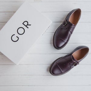 K&ouml;nnte beinhalten: Ein Paar dunkelbraune Lederschuhe mit je einem Schnallenriemen. Die Schuhe liegen auf einer wei&szlig;en Holzoberfl&auml;che. Das Wort "GOR" ist auf einem wei&szlig;en Karton im Hintergrund aufgedruckt.