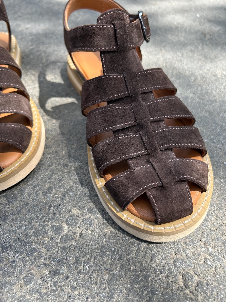 Pu&ograve; includere: Primo piano di sandali in camoscio marrone. I sandali presentano un design a gabbia con pi&ugrave; cinturini e una chiusura con fibbia. Le suole sono beige con un sottopiede in pelle marrone. Sandali comodi ed eleganti.