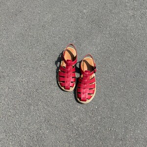 Puede incluir: Un par de sandalias de cuero rojo con un dise&ntilde;o tejido. Las sandalias tienen una plantilla beige y una peque&ntilde;a correa en el tobillo. Las suelas son de color beige claro. Las sandalias est&aacute;n sobre una superficie gris.