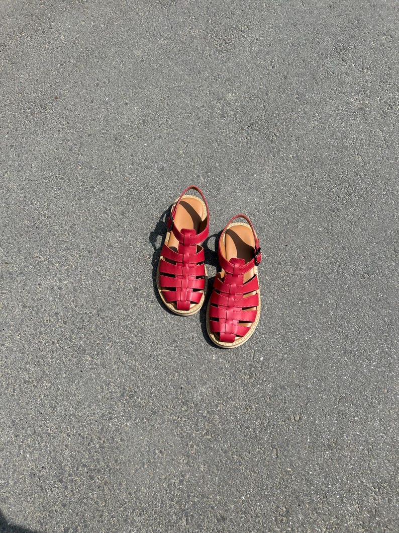 Puede incluir: Un par de sandalias de cuero rojo con un dise&ntilde;o tejido. Las sandalias tienen una plantilla beige y una peque&ntilde;a correa en el tobillo. Las suelas son de color beige claro. Las sandalias est&aacute;n sobre una superficie gris.