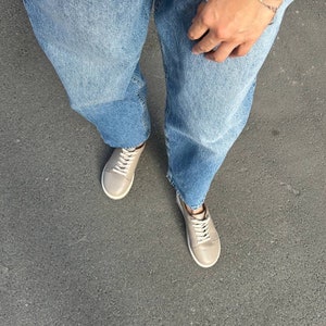 May include: A person wearing light blue denim jeans and beige leather low-top sneakers. The person is wearing a white t-shirt and a silver watch.