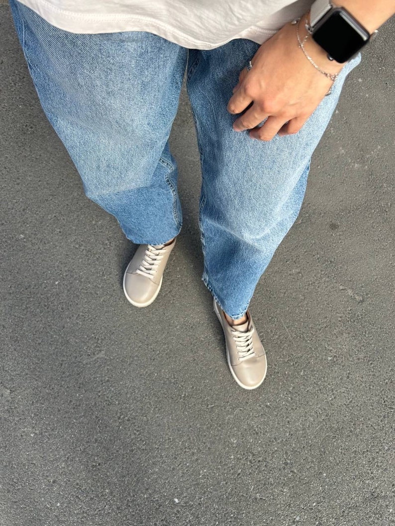 May include: A person wearing light blue denim jeans and beige leather low-top sneakers. The person is wearing a white t-shirt and a silver watch.