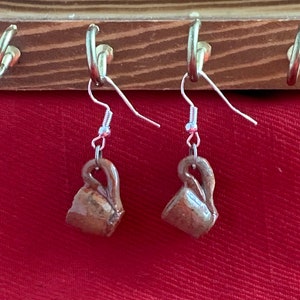 Puede incluir: Un par de pendientes de cerámica marrón con forma de pequeñas tazas, colgados de ganchos plateados. Los pendientes se exhiben sobre un fondo de tela roja y un estante de madera con ganchos plateados.