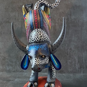 May include: A hand-carved wooden bull figurine with black, white, blue, red, and yellow paint. The bull is standing on its hind legs with its front legs raised. The bull has a long, curled tail and large, blue ears. The bull is standing on a red base with yellow dots.