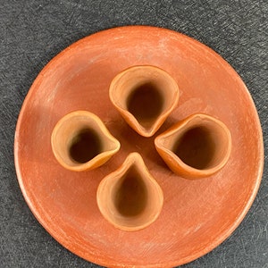 Op de afbeelding: Een set van vier terracotta shotglazen, gerangschikt in een cirkelvormige opstelling op een terracotta bord. De glazen zijn allemaal even groot en hebben dezelfde vorm, en ze hebben een gladde, afgeronde afwerking.