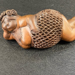 May include: A wooden sculpture of a reclining woman with a floral crown, intricate carvings, and a detailed skirt. The sculpture is made of a light brown wood and has a smooth, polished finish.