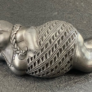 May include: A silver-colored ceramic sculpture of a reclining woman with a detailed patterned dress. The sculpture has a realistic, stylized appearance.