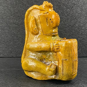 May include: A yellow ceramic sculpture of a seated figure with a large head and a detailed face. The figure is holding a rectangular object in its hands.