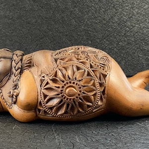 May include: A wooden sculpture of a reclining figure with intricate carvings. The figure has a braided hairstyle and is adorned with a necklace. The body features detailed floral and geometric patterns. The sculpture is a warm brown color.