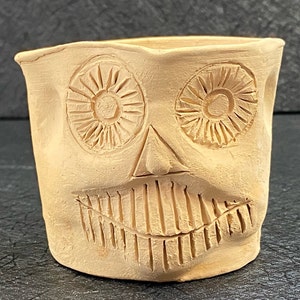 May include: A beige ceramic mug with a skull face design. The mug has two large eyes with a circular pattern and a mouth with a series of lines.