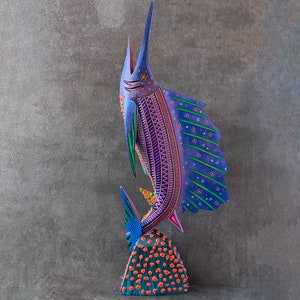 May include: A colorful wooden sculpture of a marlin fish with purple, blue, green, and pink paint. The fish is standing on a base with orange and blue paint.