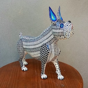 May include: A hand-carved wooden dog sculpture with black and white geometric patterns and blue accents. The dog is standing on a wooden surface.