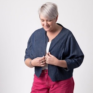 Pode incluir: Casaco azul escuro com textura e design aberto. O casaco tem mangas três-quartos e é usado sobre uma blusa branca. A pessoa na imagem está usando calças rosa. O fundo é branco.