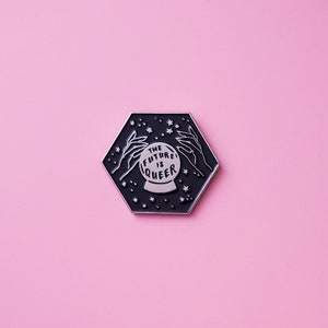 Peut inclure: Épinglette hexagonale noire avec un contour argenté. L'épingle représente un globe de cristal avec le texte "THE FUTURE IS QUEER" à l'intérieur. Le globe de cristal est entouré de deux mains et d'étoiles.