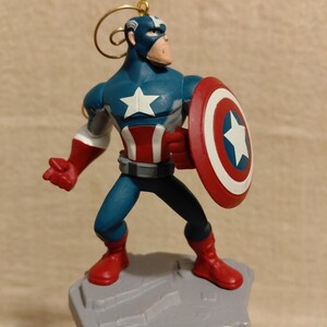 May include: A Captain America figurine in a heroic pose, holding his iconic shield. The figurine is predominantly blue, red, and white, with a gold loop for hanging. It stands on a gray, rocky base, perfect for Marvel fans.