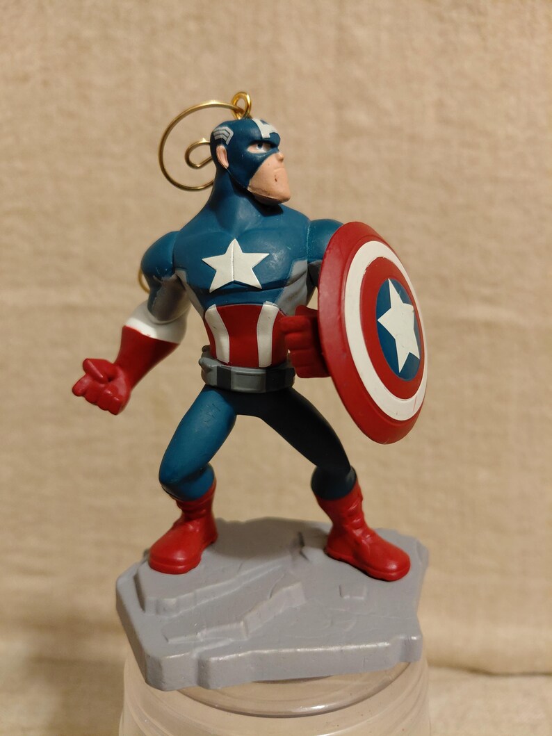 May include: A Captain America figurine in a heroic pose, holding his iconic shield. The figurine is predominantly blue, red, and white, with a gold loop for hanging. It stands on a gray, rocky base, perfect for Marvel fans.