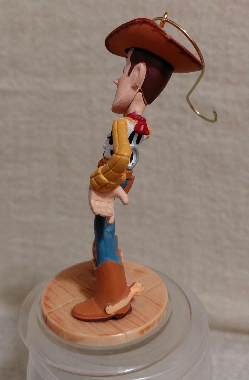 May include: A figurine of the character Woody from the animated film Toy Story. Woody is wearing a brown cowboy hat, a yellow shirt, blue jeans, and brown boots. The figurine is standing on a wooden base and has a gold wire loop for hanging.
