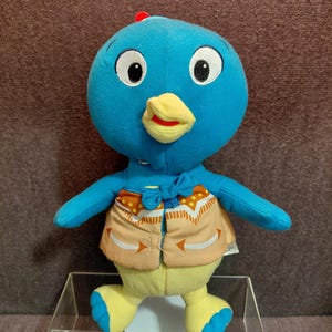 Puede incluir: Un juguete de peluche con forma de pájaro con cabeza azul, pico amarillo y ojos grandes. Lleva un chaleco color beige con un estampado geométrico y tiene brazos y piernas azules. Una pequeña cresta roja está en su cabeza.