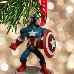 May include: A Captain America Christmas ornament. The figurine is in a heroic pose, holding his shield. The ornament is blue, red, and white, with a star on the shield and chest. It hangs from a gold wire and a red ribbon that says "MERRY CHRISTMAS".